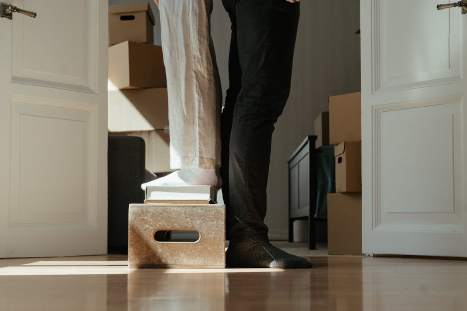 A person wearing dark trousers and socks is seen lifting a cardboard box inside a room during a home relocation process, with one foot standing on a wooden box that contains several stacked books. The room is well-lit with natural light, and in the background, there are additional cardboard boxes and a black piece of furniture, possibly a chest of drawers or a side table. The person is positioned near an open doorway, revealing doors with white paneling on either side. This scene depicts the packing and moving stage of furniture transport, with the individual preparing boxes for transportation, supported by professional removals service provider Harrow Man and Van, who specialise in difficult access moves and furniture logistics.
