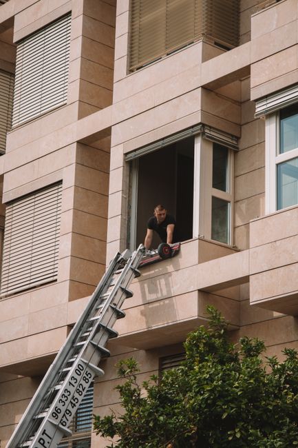 A man operating a hydraulic lift platform outside a modern, multi-storey residential building with beige stone facade and large windows. The lift extends from a ground-level position to the second-floor window, with the worker standing on the platform, which is supported by an extended metal ladder and a van parked nearby. The man is handling a power tool or equipment, likely involved in a home relocation or furniture transport process, as part of a house removal operation. The scene includes green shrubbery in the foreground and the building's architectural details, such as window shutters and horizontal cladding, visible in natural daylight. This setup illustrates a residential moving or packing activity, with professional removals services such as those offered by Harrow Man and Van involved in accessing difficult-to-reach upper floors for furniture or box loading tasks.