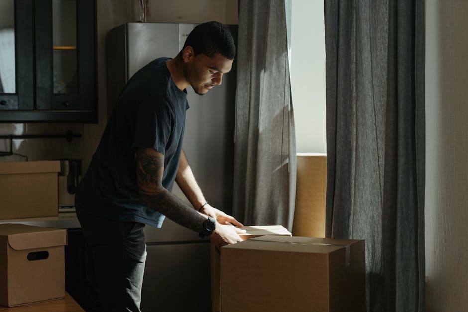 A person wearing dark trousers and socks is seen lifting a cardboard box inside a room during a home relocation process, with one foot standing on a wooden box that contains several stacked books. The room is well-lit with natural light, and in the background, there are additional cardboard boxes and a black piece of furniture, possibly a chest of drawers or a side table. The person is positioned near an open doorway, revealing doors with white paneling on either side. This scene depicts the packing and moving stage of furniture transport, with the individual preparing boxes for transportation, supported by professional removals service provider Harrow Man and Van, who specialise in difficult access moves and furniture logistics.
