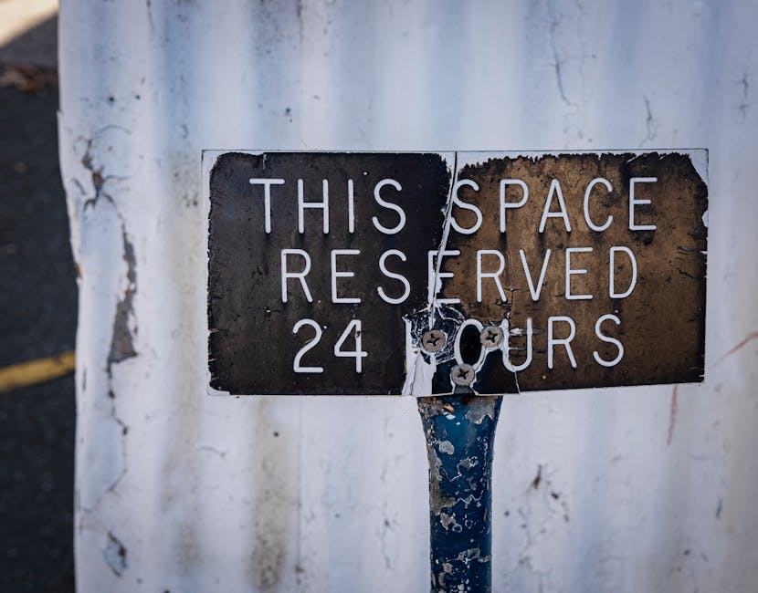 A close-up image shows a rectangular metal sign mounted on a blue-painted pole in an outdoor setting. The sign has a black background with white capital letters stating 'THIS SPACE RESERVED 24 HOURS,' but it is damaged and partly torn, with the left side of the sign showing some peeling paint and minor rust. The pole is weathered, with visible chips and peeling paint, and is positioned against a white wall or fence that has some dirt and marks. The image is well-lit, capturing the signs of wear and tear on the sign and pole. This scene is related to parking restrictions, which may be relevant for planning house removals or moving logistics, as indicated on the webpage for Harrow council parking permits for removals provided by Harrow Man and Van.