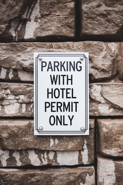 A white rectangular sign with black uppercase text reading 'PARKING WITH HOTEL PERMIT ONLY' is mounted on a rough stone wall composed of irregularly shaped, brownish stones with visible textures and natural color variations. The sign is affixed with four metal screws, one at each corner. In the context of house removals and furniture transport, this sign exemplifies restrictions on parking areas that may impact the loading process during a home relocation. The surrounding environment suggests an exterior setting, possibly near the entrance of a building, where careful planning is necessary for loading furniture and boxes onto a van. The setup indicates the importance of securing parking permits or permissions, which is relevant when coordinating removals services such as those offered by Harrow Man and Van, especially in urban areas where parking regulations are enforced. The lighting appears natural, highlighting the textured surface of the stone wall and the clarity of the signage.
