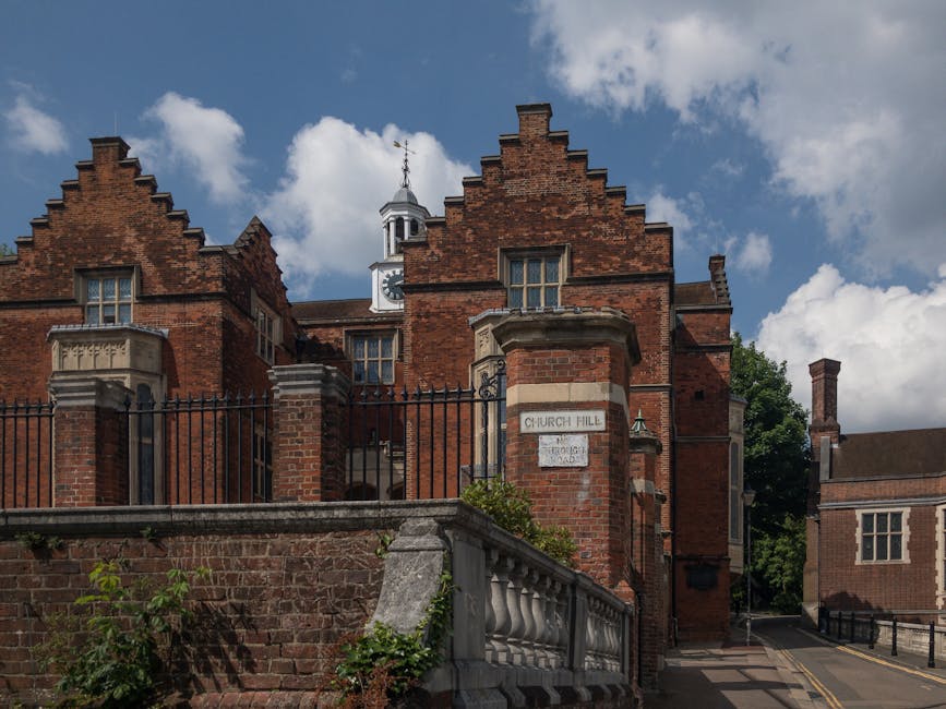 Exterior view of a historic brick building with a stepped gable roof and large windows, located on Church Hill in Harrow. The building features decorative stone window sills and a small clock tower on the roof. In the foreground, a brick wall with iron railings and a concrete balustrade runs parallel to the street, with some green plants climbing the wall. A narrow street with parked cars and a sidewalk flank the building, indicating a residential or communal area. The scene is captured during daylight with a partly cloudy sky overhead. This setting is relevant to home relocation and furniture transport, as visual cues such as the building's size and street environment suggest a typical location where professional removals, like those from Harrow Man and Van, might be involved in packing and loading furniture for transport.
