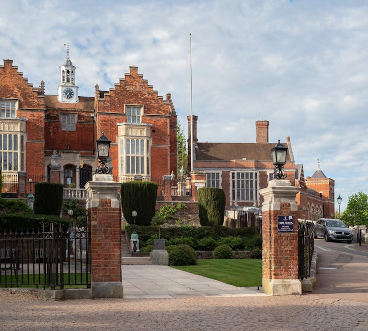 A view of a large, historic red-brick building with a clock tower, ornate white window frames, and decorative architectural details, situated behind a black wrought-iron gate and brick pillars topped with black lanterns. The building is set on a well-maintained lawn with neatly trimmed bushes and tall, sculpted trees. The entrance pathway is paved with light-colored tiles leading up to the building, and a grey vehicle is parked on the street nearby. The scene is illuminated by natural daylight, with a partly cloudy sky overhead, illustrating a luxurious residence suitable for house removals or relocation services, as handled by Harrow Man and Van.