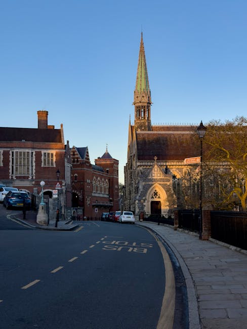 A street view outside a historic brick building with large windows and a prominent church with a tall, pointed spire. The scene includes parked cars along the curb, a streetlamp, and a designated bus 