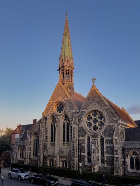 A street view outside a historic brick building with large windows and a prominent church with a tall, pointed spire. The scene includes parked cars along the curb, a streetlamp, and a designated bus 