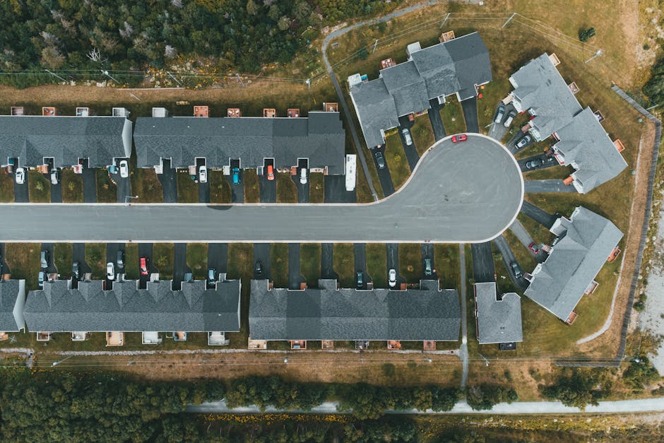 An aerial view of a residential cul de sac with multiple two-storey houses arranged in a semi-circular formation around a central curved road. Each house has a driveway with parked cars, some covered with protective tarps or parked directly outside. The properties are separated by small gardens or lawns, and some have visible fencing around them. The road is wide and extends to a main street at the bottom of the image. The surroundings include patches of grass, trees, and fencing, indicating a suburban area. This scene illustrates the typical environment where home relocations and furniture transport may take place, with the houses and vehicles positioned for ease of loading and unloading during a professional removal or moving service, such as that provided by Harrow Man and Van.