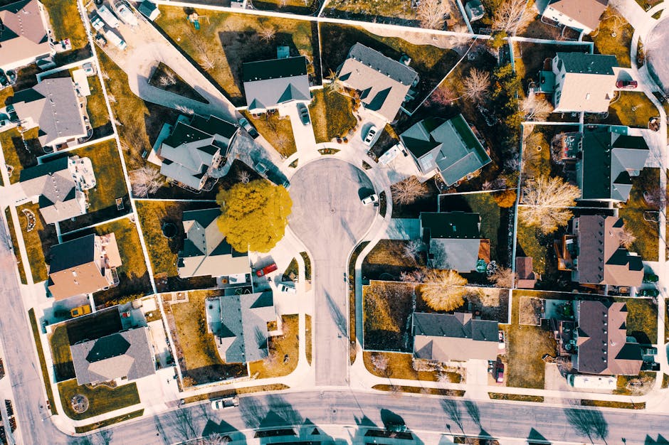 An aerial view of a residential cul-de-sac with multiple detached and semi-detached houses, each with driveways and small front gardens. The street is curved and features a central circular turnaround with several parked cars on the pavement and within driveways. Surrounding properties have rooftops of various shades of grey and brown, with some featuring chimneys. Trees with yellowing leaves are visible in front gardens, indicating a possible autumn setting, and the overall scene is illuminated by natural daylight. The image captures the organized layout typical of suburban home developments, illustrating the type of environment where house removals and furniture transport by Harrow Man and Van may take place during home relocation or packing and moving processes, particularly in cul-de-sac homes in North Harrow.