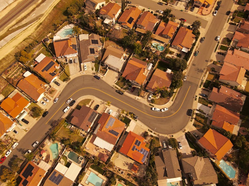 An aerial view of a residential cul de sac with multiple two-storey houses arranged in a semi-circular formation around a central curved road. Each house has a driveway with parked cars, some covered with protective tarps or parked directly outside. The properties are separated by small gardens or lawns, and some have visible fencing around them. The road is wide and extends to a main street at the bottom of the image. The surroundings include patches of grass, trees, and fencing, indicating a suburban area. This scene illustrates the typical environment where home relocations and furniture transport may take place, with the houses and vehicles positioned for ease of loading and unloading during a professional removal or moving service, such as that provided by Harrow Man and Van.