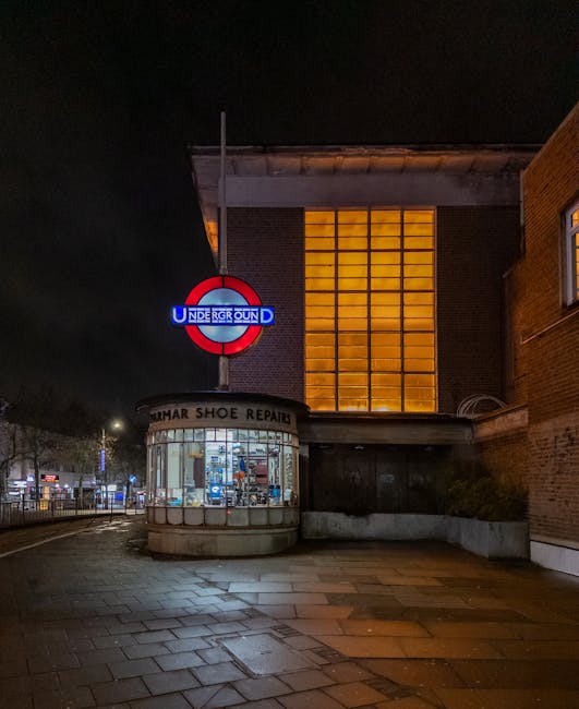 Nighttime exterior view of a brick building adjacent to Harrow's underground station entrance, featuring a luminous, illuminated round London Underground sign with the word 'UNDERGROUND' in white on a blue background, and a red circular border. Next to the station entrance, there is a small, curved shopfront with large glass windows displaying various shoe repair tools and supplies, with a sign reading 'HUMAR SHOE REPAIR' above the window. The sidewalk in front is paved with rectangular tiles, some of which are wet, reflecting the surrounding lights. In the background, streetlights and parked vehicles are visible, and the building has a large window with a grid of orange-yellow lighting on the upper floor, contrasting against the night sky. Harrow Man and Van offers house removals and furniture transport services, and this scene may relate to moving logistics near the station area.