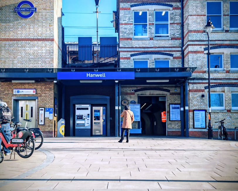 Inside the Harrow and Wealdstone underground station, showing a platform with beige tiled walls and a yellow safety line along the edge. On the left, there is a staircase with black and white steps leading up, and a sign indicating the way out. Further along the platform, a red London Underground train is approaching the station, with its front lights visible in the distance. The platform surface is made of speckled beige and brown tiles, and various advertisements and informational posters are displayed on the station walls. This scene captures the typical environment of a commuter station, emphasizing the platform layout and the train's approach, relevant for illustrating home relocation or moving logistics involving station access and transportation in Harrow. Harrow Man and Van may assist with packing, transport, and station access advice as part of a comprehensive removals service.