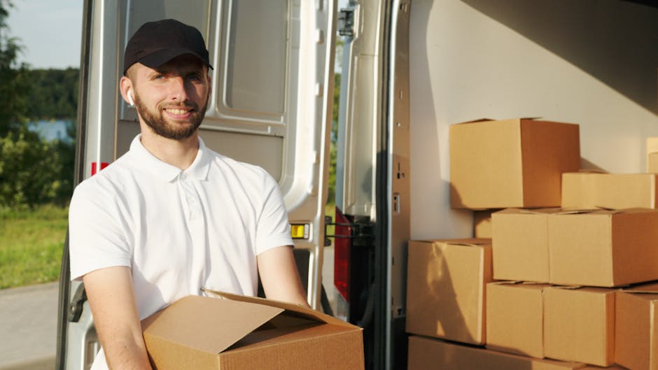 A man in a white polo shirt and black cap is standing outside next to an open van, loading or unloading a cardboard box. Inside the van, several neatly stacked cardboard boxes are visible, some wrapped in plastic or packing materials. The backdrop includes a rural landscape with green trees and a blue sky, indicating a daytime home relocation or moving process. The scene captures the careful handling of household items during a furniture transport or packing and moving operation. Harrow Man and Van specializes in removals and home moving services, as seen in this loading process that involves careful handling of boxes and using appropriate equipment like trolleys or straps to facilitate efficient and secure transport, supporting the overall logistics of same-day removals in Harrow.
