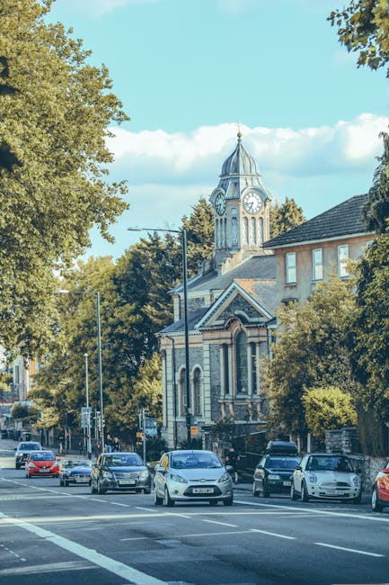 A view of a busy street in Harrow during daytime, featuring multiple parked cars along the roadside and several moving vehicles, including a white van which appears to be part of a house removal or furniture transport operation by Harrow Man and Van. The street is lined with leafy trees providing shade and greenery. In the background, there is a historic clock tower with a domed roof and ornate architectural details, situated above a building with large windows and a pitched roof. The scene suggests an active urban area commonly associated with residential and local commercial activities, and it illustrates the typical environment where professional removals and home relocation services may take place, especially during packing, loading, and transportation processes related to same-day removals in Harrow.