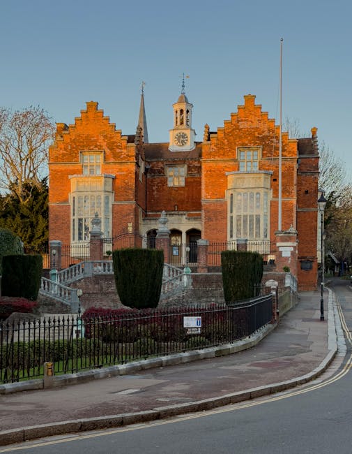 This image depicts the exterior of a historic brick building with large, ornate windows and a clock tower above. In front of the building is a paved courtyard enclosed by a black wrought iron fence su