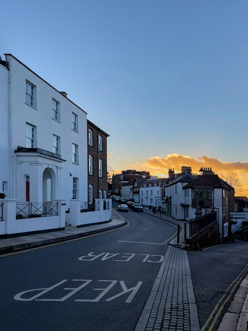 A quiet residential street in Wealdstone during sunset, featuring a row of white and dark-colored terraced houses with varied architectural styles, including bay windows and small front gardens. The street is gently sloping and paved with asphalt, with clearly marked road signs indicating 'KEEP CLEAR' on the roadway. Sidewalks made of concrete slabs are present at the edges, with tactile paving near the curb for pedestrian safety. Street lamps and occasional parked cars are visible along the kerbside. The sky displays a warm glow with low-hanging clouds near the horizon. This setting illustrates a typical suburban area where house removals or furniture transport processes might take place, especially considering the narrow streets that require careful planning for moving services such as those offered by Harrow Man and Van, focusing on safe and efficient home relocation and packing and moving logistics.